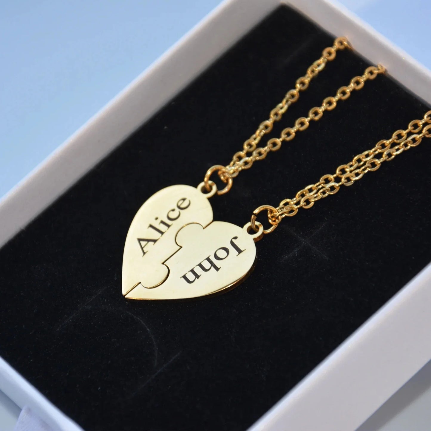 Gold heart-shaped puzzle piece pendant with engraved names on a black cushion in a white box.